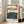May include: Framed vintage Japanese woodblock print poster leaning against white wall next to beige sofa in bright living room with large windows. The artwork depicts Mount Fuji above a bustling blue-toned street scene with many small figures, traditional buildings, and colorful sky. Print has cream matting and light wood frame, shows classic ukiyo-e art style with red cartouche in upper corner and descriptive text below image.