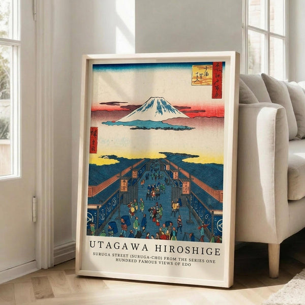 May include: Framed vintage Japanese woodblock print poster leaning against white wall next to beige sofa in bright living room with large windows. The artwork depicts Mount Fuji above a bustling blue-toned street scene with many small figures, traditional buildings, and colorful sky. Print has cream matting and light wood frame, shows classic ukiyo-e art style with red cartouche in upper corner and descriptive text below image.