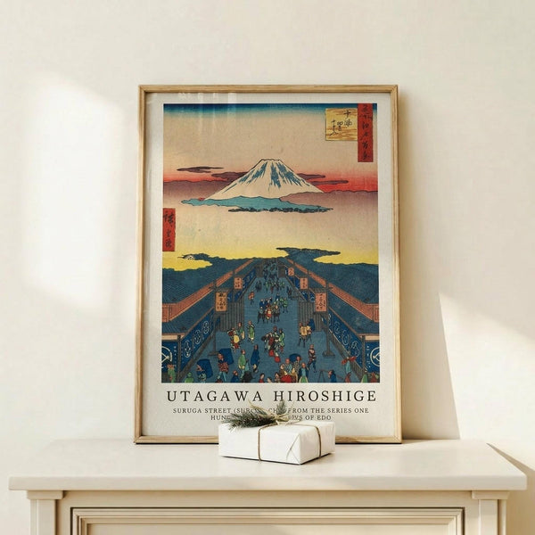 May include: Framed Japanese woodblock print featuring Mount Fuji in the distance above a bustling blue-roofed street scene with small figures in traditional dress. The artwork displays classic ukiyo-e style with muted earth tones, yellow sky, and red cartouche with Japanese characters. Light wood frame sits on pale wooden surface beside small white wrapped gift with natural twine against cream wall.