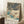 May include: Framed Japanese woodblock print featuring a snowy winter landscape with snow-covered pine trees, a blue stream, stone lantern, and footpath through a garden. The artwork displays traditional ukiyo-e style with muted blues, whites, and earth tones. Print is in light wood frame, leaning against white wall corner next to wooden side table with white coffee cup, positioned on textured white rug near beige fabric.