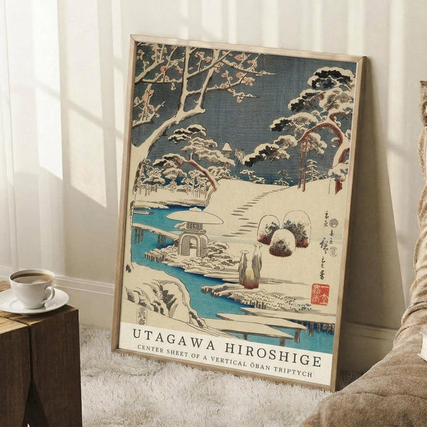 May include: Framed Japanese woodblock print featuring a snowy winter landscape with snow-covered pine trees, a blue stream, stone lantern, and footpath through a garden. The artwork displays traditional ukiyo-e style with muted blues, whites, and earth tones. Print is in light wood frame, leaning against white wall corner next to wooden side table with white coffee cup, positioned on textured white rug near beige fabric.