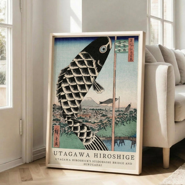 May include: A framed Japanese woodblock print featuring a large black and white scaled carp or koi fish against a blue sky, with Mount Fuji and traditional buildings visible in the background landscape. The artwork displays traditional ukiyo-e style with Japanese text characters in red cartouches. The light wood frame leans against a white wall next to a beige upholstered sofa in a bright, modern living room with large windows.