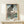 May include: Japanese woodblock print poster in light wood frame showing large black koi fish with detailed scales against blue-green landscape background featuring Mount Fuji. Print displays traditional ukiyo-e art style with Japanese text. Frame sits on light wood surface next to small white gift box with green sprig. Cream colored wall background creates minimalist display setting.