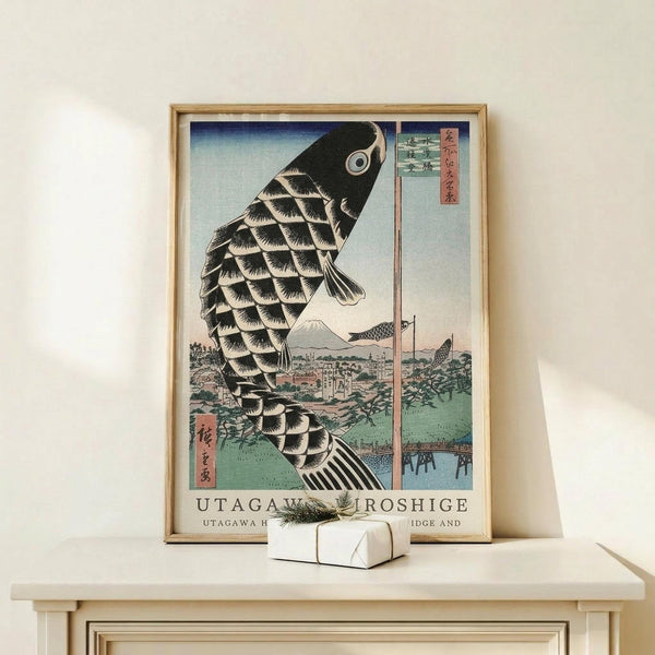 May include: Japanese woodblock print poster in light wood frame showing large black koi fish with detailed scales against blue-green landscape background featuring Mount Fuji. Print displays traditional ukiyo-e art style with Japanese text. Frame sits on light wood surface next to small white gift box with green sprig. Cream colored wall background creates minimalist display setting.