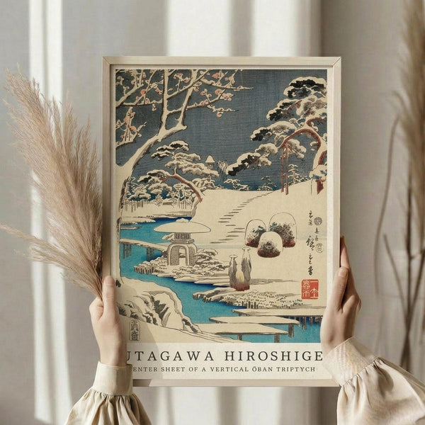 May include: Hands holding a framed Japanese woodblock print showing a snowy winter landscape with snow-covered trees, a stone lantern, winding path, and blue stream. The artwork features traditional ukiyo-e style with muted colors against cream background. Person wears beige clothing with gathered sleeves, dried pampas grass visible on left side.