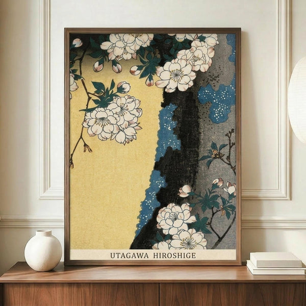 May include: Framed Japanese woodblock print featuring cherry blossoms with white petals and green leaves against golden yellow and blue background, displayed on wooden sideboard next to white ceramic vase and books in elegant room with cream walls and white molding.