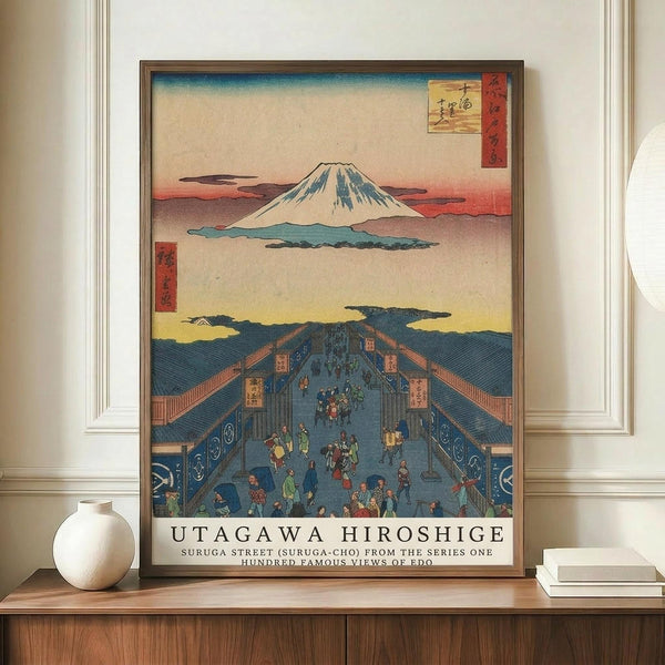 May include: Japanese woodblock print poster displayed on wooden sideboard featuring Mount Fuji in background and bustling Edo period street scene with people walking through covered marketplace. Blue-roofed buildings line the street with colorful banners and signs. Print shows traditional ukiyo-e art style with muted earth tones and blue accents, framed in dark wood frame. White ceramic vase and books placed nearby on furniture.
