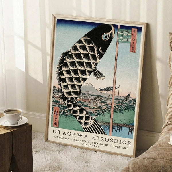 May include: Framed vintage Japanese woodblock print featuring a large black and white scaled carp kite against blue sky, with Mount Fuji and traditional village landscape below. The artwork displays red Japanese text columns and sits in a light wood frame on white carpet beside a coffee cup on wooden block and beige fabric chair in bright natural lighting.