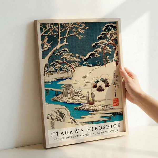 Japanese woodblock print showing a snow-covered winter garden scene with traditional architecture, stone lanterns, and pine trees heavy with snow. Features a bright turquoise stream winding through white snowy landscape, with small figures walking along paths. The artwork displays classic ukiyo-e style with bold colors against a dark teal sky. Presented in a light wooden frame, held by a hand against a white wall, showing the print's medium size and museum-quality presentation.
