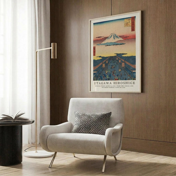 May include: Framed Japanese woodblock print poster featuring Mount Fuji and colorful landscape scenes displayed on wood-paneled wall above modern beige armchair with geometric pillow. Brass floor lamp with white base stands nearby next to dark round side table with open book. Neutral tones throughout minimalist interior setting.