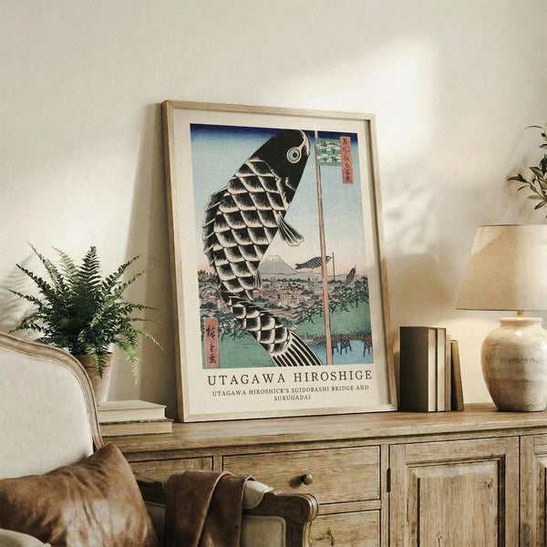 May include: Framed Japanese woodblock print featuring a large black carp kite with detailed scales floating above a landscape with Mount Fuji in the background. The artwork displays traditional ukiyo-e style with blue sky gradients and includes Japanese text. Print sits on rustic wooden sideboard next to fern plant, ceramic vase, and stacked books in warmly lit living room setting with cream upholstered chair visible.