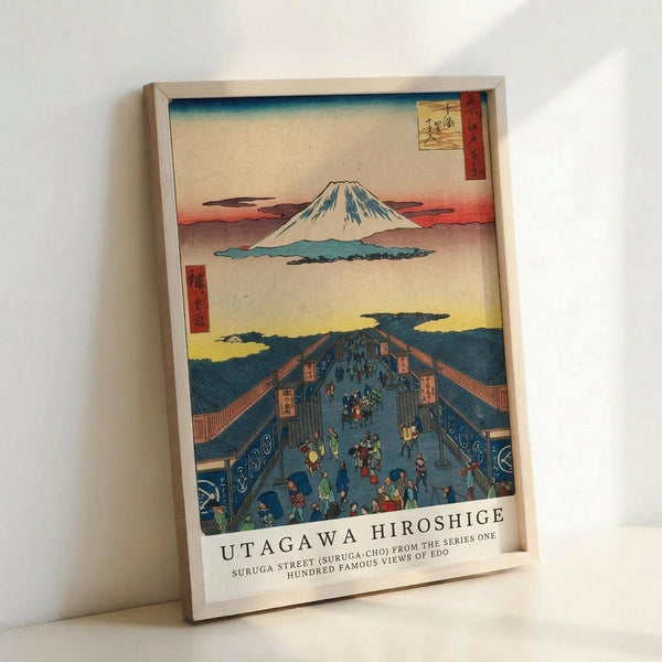 A framed Japanese woodblock print reproduction showing Mount Fuji in the background with snow-capped peak against a gradient sky of blue, pink and yellow. The foreground depicts a busy blue-roofed street scene with many small colorful figures walking along what appears to be a covered marketplace or shopping district. Red text cartouches with Japanese characters appear in the upper corners. The artwork is displayed in a light wooden frame against a white wall, showing the classic ukiyo-e art sty