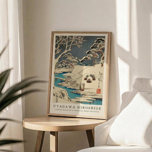 May include: Framed vintage Japanese woodblock print showing snowy winter landscape with snow-covered trees, blue stream, small figures, and traditional buildings. Print displays in thin wooden frame on round wooden side table next to beige upholstered chair with plant leaves visible in corner. Soft natural lighting creates shadows on neutral wall.