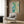 May include: Abstract watercolor artwork in turquoise, teal and green tones with organic flowing shapes and ink blot effects, displayed in light wood frame on warm brown wood paneled wall. Modern beige armchair with geometric patterned pillow sits below, brass floor lamp and side table nearby in contemporary living room setting.