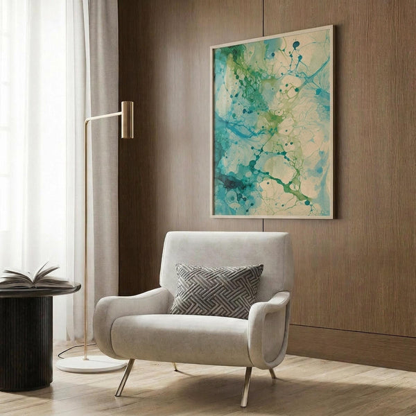 May include: Abstract watercolor artwork in turquoise, teal and green tones with organic flowing shapes and ink blot effects, displayed in light wood frame on warm brown wood paneled wall. Modern beige armchair with geometric patterned pillow sits below, brass floor lamp and side table nearby in contemporary living room setting.