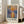 May include: Framed poster print of Impressionist-style harbor scene at sunrise with sailing ships silhouetted against orange and yellow sky reflecting on blue water, displayed in light wood frame on floor beside beige sofa in bright modern living room with white walls and large windows