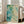 May include: Large framed abstract art print leaning against white wall beside beige upholstered chair. Artwork features flowing teal, turquoise, and green watercolor-style ink blots with organic branching patterns and scattered dots on cream background. Natural wood frame, bright sunlit room with hardwood floors and windows.