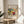 May include: Framed abstract art print in thin gold frame displayed on round wooden side table next to beige upholstered chair. Colorful expressionist painting features bold brushstrokes in yellow, red, blue, and green with abstract forms. White mat with black text identifying artwork and artist. Soft natural lighting creates shadows on neutral wall. Green plant leaves visible in corner of modern, minimalist room setting.