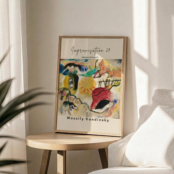 May include: Framed abstract art print in thin gold frame displayed on round wooden side table next to beige upholstered chair. Colorful expressionist painting features bold brushstrokes in yellow, red, blue, and green with abstract forms. White mat with black text identifying artwork and artist. Soft natural lighting creates shadows on neutral wall. Green plant leaves visible in corner of modern, minimalist room setting.
