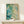 May include: Abstract watercolor artwork in wooden frame displayed on light wood console table against cream wall. Fluid organic shapes in turquoise, teal, sage green and cream tones with connected dots and flowing lines throughout. Small wrapped gift with natural sprig placed in front of vertical rectangular frame.