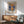 May include: Framed poster of impressionist harbor scene at sunrise with sailing ships silhouetted against orange and blue sky, water reflecting golden sunlight. Wood-framed print hangs above mid-century modern credenza with white ceramic lamp. Sage green upholstered chair with wooden arms sits nearby. Warm natural light creates shadows across white wall in contemporary living room setting.