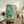 May include: Abstract watercolor artwork in turquoise, teal, and green tones with organic flowing shapes and scattered dots, displayed in light wood frame on rustic wooden sideboard. Room scene shows fern plant, stacked books, ceramic vase, and beige upholstered chair in warm, naturally-lit interior setting.