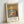 May include: Framed art print leaning against white wall showing impressionist-style beach sunset scene with golden sky, blue ocean waves, sandy shore, and solitary figure walking. Warm orange and yellow tones reflect on water. Light wooden frame with white matting. Text reads "MONET" and "ART CONTEMPORAIN INSPIRÉ PAR UN MAÎTRE ARTISTIQUE" below image.