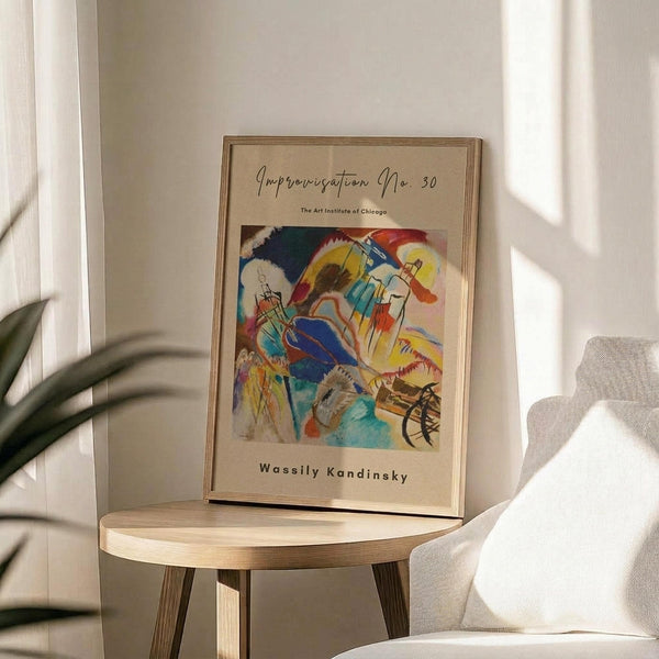 May include: Framed art print of Wassily Kandinsky's abstract painting featuring vibrant swirls of blue, yellow, red and green with geometric shapes and flowing lines. The beige matted poster sits on a light wood round side table next to a cream upholstered chair, with plant leaves visible in soft natural lighting against white walls.