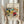 May include: Person in beige ruffled sleeves holding framed abstract art print featuring vibrant swirling colors in yellow, red, blue, and green with flowing organic shapes. Natural pampas grass visible on left side. Light wood frame against soft neutral curtained background creates warm, minimalist home decor setting.
