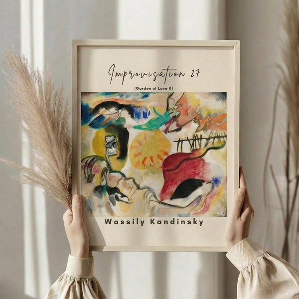 May include: Person in beige ruffled sleeves holding framed abstract art print featuring vibrant swirling colors in yellow, red, blue, and green with flowing organic shapes. Natural pampas grass visible on left side. Light wood frame against soft neutral curtained background creates warm, minimalist home decor setting.
