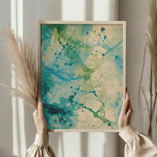 May include: Person in cream sweater holding framed abstract artwork with flowing teal, turquoise and sage green ink blot patterns on cream background. Light wood frame, dried pampas grass visible to left, soft natural lighting near window with sheer curtains.