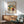 May include: Framed abstract art print displayed on white wall above wooden sideboard with table lamp. Green upholstered mid-century armchair with wooden arms sits nearby. The colorful abstract artwork features vibrant blues, yellows, reds and greens with flowing organic shapes. Natural sunlight creates dramatic shadows across the minimalist room setting with light wood furniture and neutral tones.