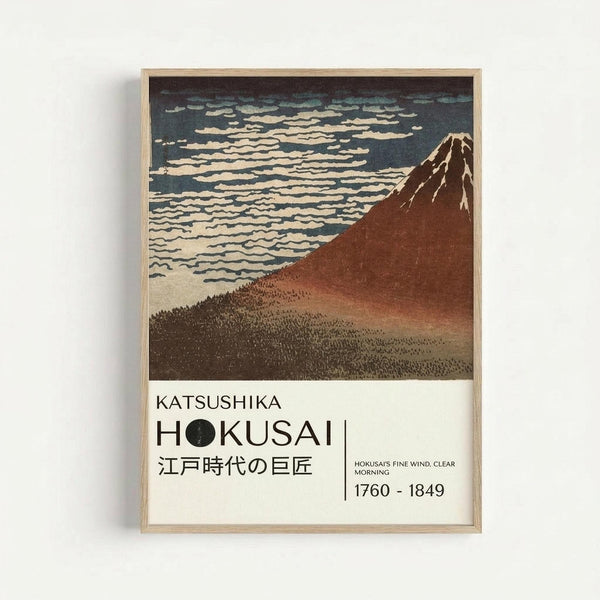 May include: Framed Japanese woodblock print poster showing Mount Fuji in reddish-brown tones against a blue sky with white clouds, displayed on a white wall. The artwork features traditional ukiyo-e style with Japanese text below the mountain scene. Light wooden frame surrounds the print with museum-style labeling at bottom including artist name Hokusai and dates 1760-1849.
