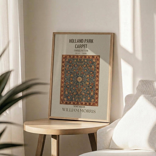 May include: Framed vintage-style carpet design poster displayed on light wood round side table next to beige upholstered chair. Print features ornate floral pattern in deep blues, oranges and golds with decorative border, housed in thin gold frame. Soft natural lighting creates shadows across neutral wall, with green plant leaves visible in corner.