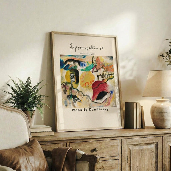 May include: Framed abstract art print displayed on rustic wooden sideboard in neutral-toned living room. Colorful Kandinsky-style artwork features bold brushstrokes in blues, yellows, reds, and greens with organic flowing forms. Light wood frame complements natural decor including fern plant, ceramic vase, stacked books, and cream upholstered furniture with brown leather cushion.