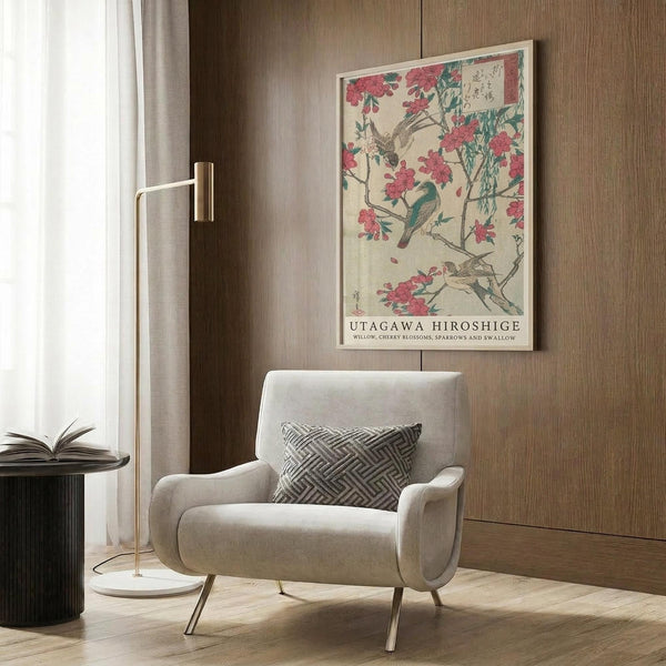 May include: Japanese woodblock print poster featuring birds perched on flowering cherry blossom branches in pink and green tones, displayed in light wooden frame on wood paneled wall above cream upholstered armchair with geometric patterned pillow, brass floor lamp and dark round side table with open book nearby