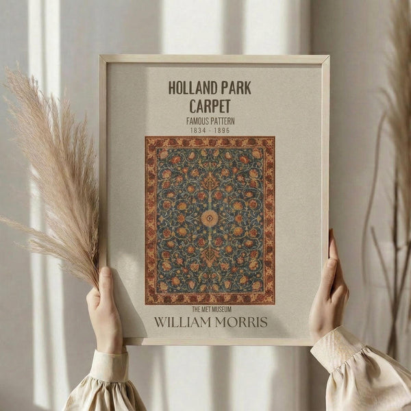 May include: Hands holding a framed vintage carpet design print featuring intricate floral patterns in deep blues, oranges, and golds with ornate border. Light wood frame against neutral beige mat. Person wearing cream-colored blouse with gathered sleeves holds dried pampas grass. Soft natural lighting creates gentle shadows on white curtained background.