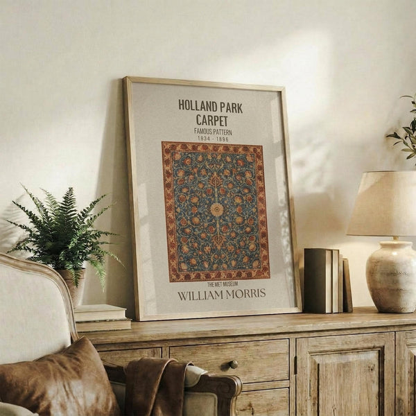 May include: Framed vintage-style museum poster featuring an ornate carpet with intricate blue and orange floral patterns displayed on a rustic wooden sideboard. The print shows William Morris design from 1834-1896. Scene includes fern plant, stacked books, ceramic vase, and cream upholstered chair in warm, naturally-lit living room setting with neutral walls.