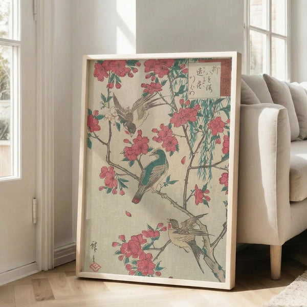 May include: Framed Japanese woodblock print featuring pink cherry blossoms, green willow branches, and colorful birds on cream background with Japanese text, displayed in light wood frame leaning against white wall next to beige upholstered chair in bright living room with large windows.