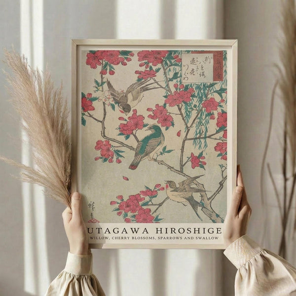 May include: Person in cream sweater holding framed Japanese woodblock print showing pink cherry blossoms, gray and teal birds, and willow branches on beige background. Light wood frame, dried wheat stalks visible, soft natural lighting against white curtained window.