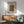 May include: Framed vintage carpet pattern print hanging above mid-century modern wooden sideboard with white ceramic lamp. Green upholstered armchair with wooden frame sits nearby. Poster features ornate floral carpet design in blues, oranges and earth tones with "Holland Park Carpet" and "William Morris" text. Natural sunlight creates shadows across white wall and warm wood furniture in minimalist living room setting.