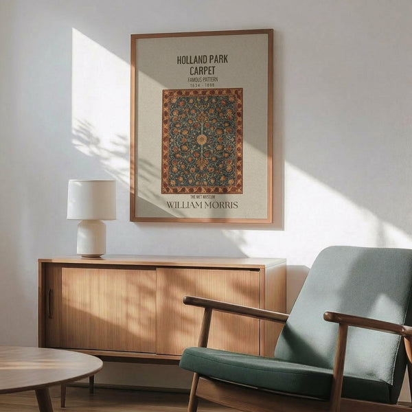May include: Framed vintage carpet pattern print hanging above mid-century modern wooden sideboard with white ceramic lamp. Green upholstered armchair with wooden frame sits nearby. Poster features ornate floral carpet design in blues, oranges and earth tones with "Holland Park Carpet" and "William Morris" text. Natural sunlight creates shadows across white wall and warm wood furniture in minimalist living room setting.