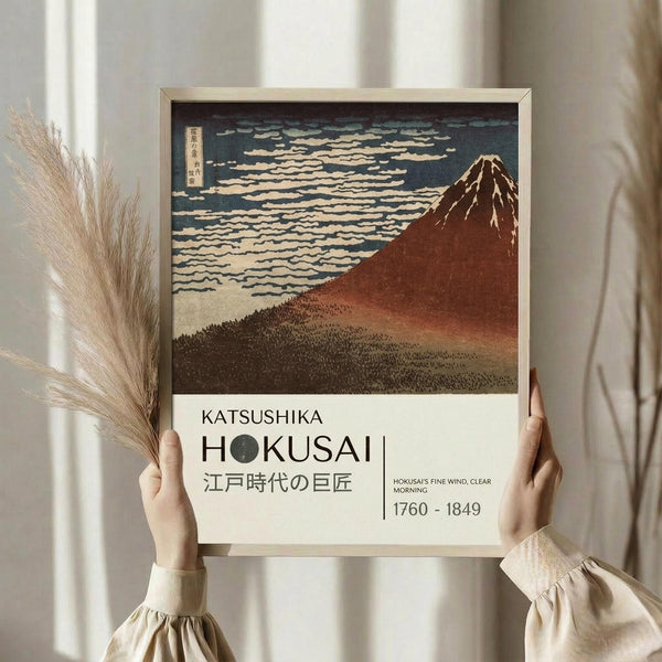 May include: Person in beige ruffled sleeves holding framed reproduction of Japanese woodblock print featuring Mount Fuji in reddish-brown tones against cloudy sky, with white text panel below reading "KATSUSHIKA HOKUSAI" and dates "1760-1849", displayed near dried pampas grass stems against neutral curtained background