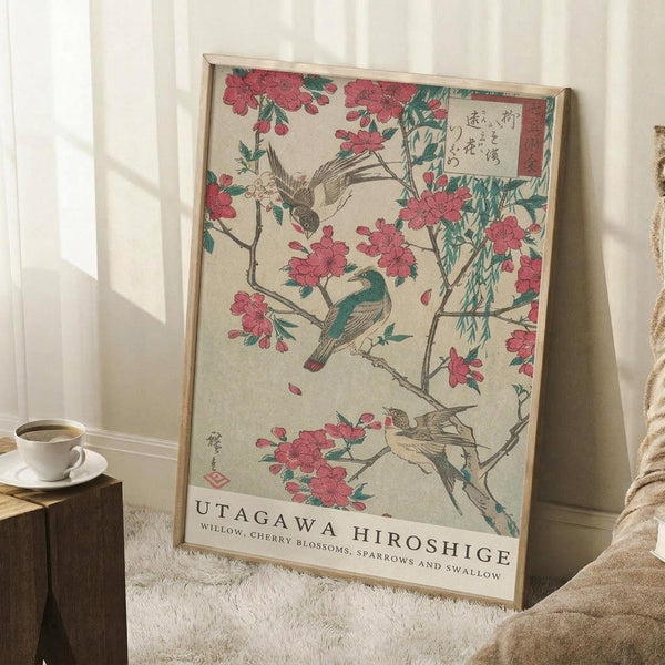 May include: A framed Japanese woodblock print featuring pink cherry blossoms on branches with three small birds - one flying at top, one perched mid-branch, and one at bottom. The artwork has muted earth tones with Japanese text in the upper right corner and "UTAGAWA HIROSHIGE" credit at bottom. The print leans against white paneled walls in a cozy corner setting with a coffee cup on a wooden block and cream blanket visible.