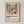 May include: Framed poster print in light wood frame mounted on cream wall. Artwork shows colorful still life with pink flowers in yellow vase, blue and green pitchers, yellow and red fruits on pink table surface. Bold shapes in Matisse-inspired style with vivid blues, pinks, oranges, and greens. White matting surrounds the square print with black text below reading "Celebrating Colors of Life" and descriptive paragraph.