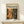 May include: Framed art poster with "Chaos and Creativity Pollock 1912-1956" text above abstract painting featuring orange, green, and black paint splatters and drips. Light wood frame displayed on pale wooden surface with small white wrapped gift box, against cream wall.