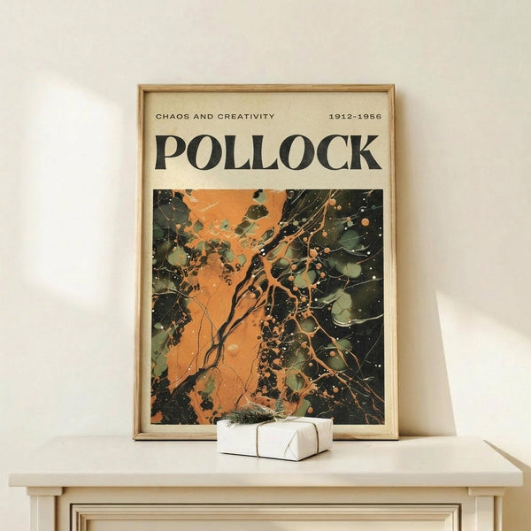 May include: Framed art poster with "Chaos and Creativity Pollock 1912-1956" text above abstract painting featuring orange, green, and black paint splatters and drips. Light wood frame displayed on pale wooden surface with small white wrapped gift box, against cream wall.