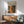 May include: Framed abstract art poster with orange and dark green paint splatters mounted above a mid-century modern wooden credenza. Scene shows mint green upholstered armchair with wooden frame, small round coffee table, and white ceramic table lamp. Warm natural light creates geometric shadows on white wall in minimalist living room setting.