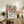 May include: Framed art print displayed on wooden console table beside fern plant and ceramic vases. Poster features colorful still life with flowers in vase, fruit, and pitcher in Matisse-inspired style with bold shapes in pink, yellow, green, and blue. Natural wood frame complements rustic furniture and neutral wall backdrop.