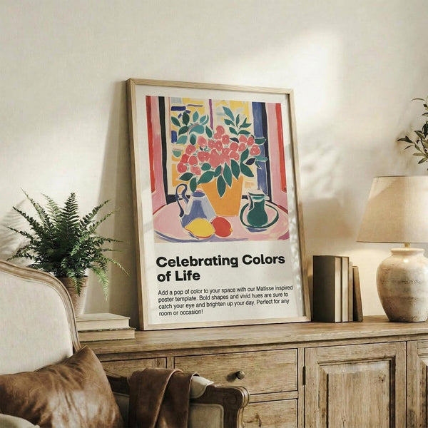 May include: Framed art print displayed on wooden console table beside fern plant and ceramic vases. Poster features colorful still life with flowers in vase, fruit, and pitcher in Matisse-inspired style with bold shapes in pink, yellow, green, and blue. Natural wood frame complements rustic furniture and neutral wall backdrop.